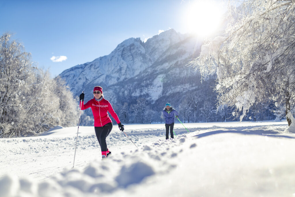 eine Frau mit pinker Jacke und ein Herr mit blauer Jacke die gerade langlaufen, schneebedeckte Winterlandschaft, links und rechts schneebedeckte Bäume, im Hintergrund Berge voller Schnee